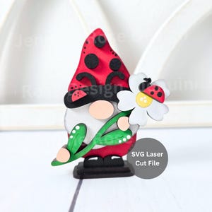 Può includere: Statuetta di gnomo in legno con cappello rosso a pois neri, che tiene una margherita bianca con un disegno a coccinella. Lo gnomo ha un viso beige e si trova su una base nera. Il testo "SVG Laser Cut File" è visibile.
