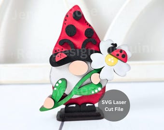 May include: A wooden gnome figurine with a red hat with black spots, holding a white daisy with a ladybug design. The gnome has a beige face and is standing on a black base. The text "SVG Laser Cut File" is visible.