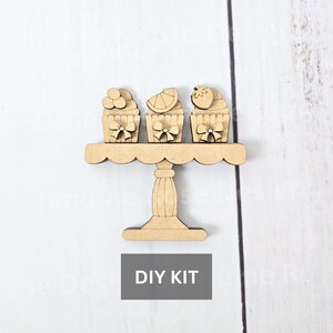 May include: A wooden DIY kit featuring a tiered cake stand with three cupcake cutouts. Each cupcake has a different topping and a bow. The text "DIY KIT" is in a gray rectangle below the stand.