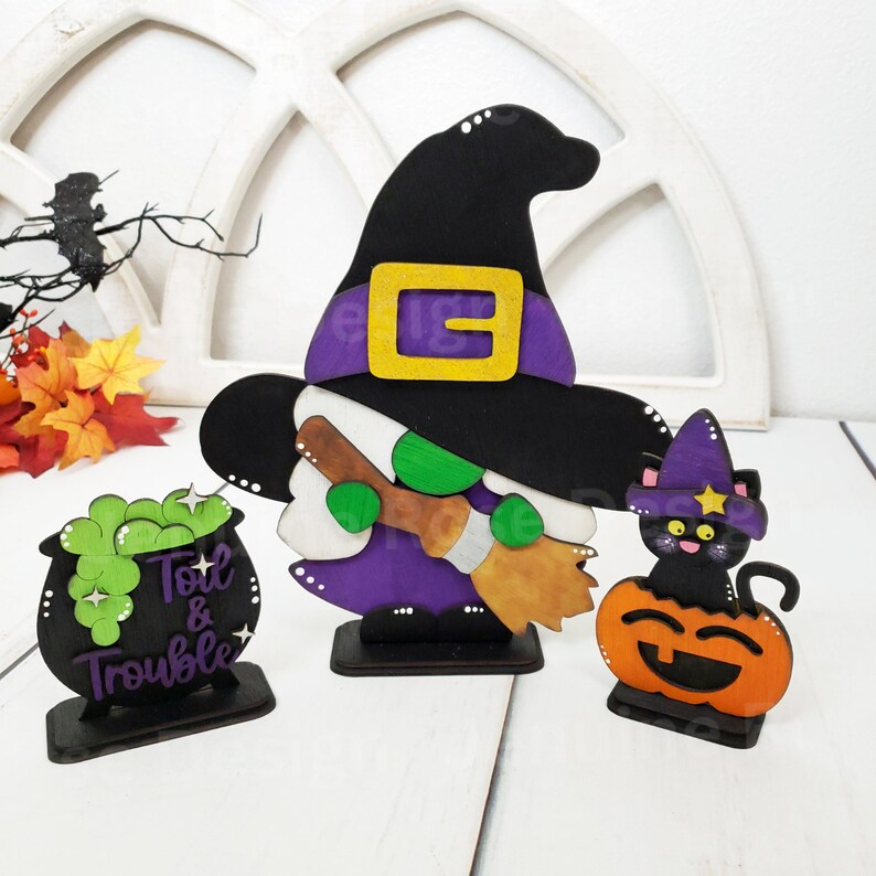 May include: A wooden witch figurine with a purple dress and black hat, holding a broom. The witch is standing on a wooden base. There is a black cat with a pumpkin head and a cauldron with the text "Toil & Trouble" on a wooden base.