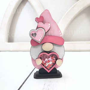 Op de afbeelding: Een roze en grijze houten gnome die een hartvormige doos vasthoudt gevuld met roze en rode glitterhartjes. De gnome draagt een roze hoed met witte stippen en heeft een witte baard.