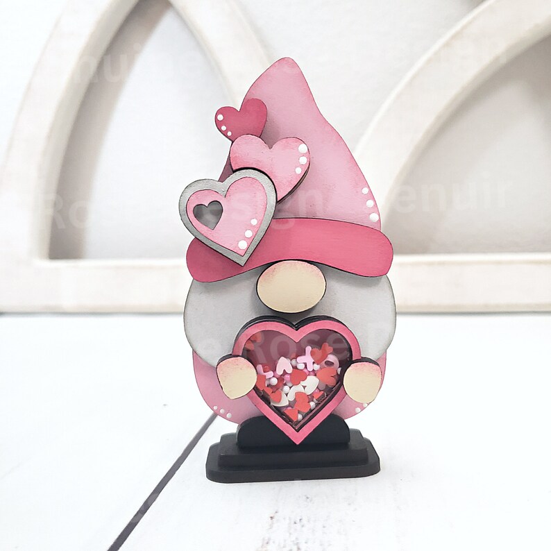 Op de afbeelding: Een roze en grijze houten gnome die een hartvormige doos vasthoudt gevuld met roze en rode glitterhartjes. De gnome draagt een roze hoed met witte stippen en heeft een witte baard.