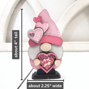 Op de afbeelding: Een roze houten gnome die een hartvormige doos vasthoudt gevuld met roze en witte confetti. De gnome draagt ​​een roze hoed met witte stippen en heeft een grijze baard. De gnome is ongeveer 10 cm hoog en 5,7 cm breed.