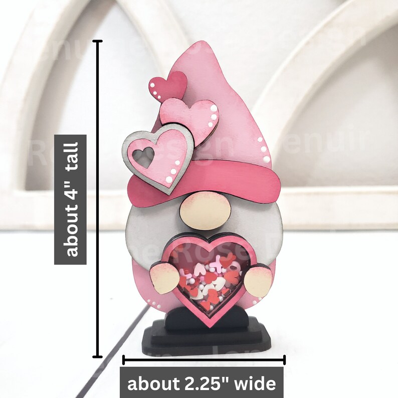 Op de afbeelding: Een roze houten gnome die een hartvormige doos vasthoudt gevuld met roze en witte confetti. De gnome draagt ​​een roze hoed met witte stippen en heeft een grijze baard. De gnome is ongeveer 10 cm hoog en 5,7 cm breed.