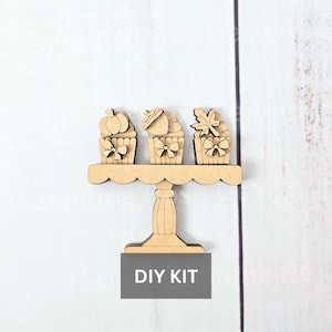 May include: A wooden DIY kit featuring three cupcake-shaped decorations with fall-themed toppers: a pumpkin, an acorn, and a leaf. Each cupcake has a bow. The cupcakes are displayed on a scalloped-edge cake stand. The text "DIY KIT" is at the bottom.