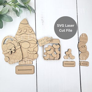 Puede incluir: Decoraciones de Pascua de madera cortadas con láser. Incluye un gnomo con una cesta, un tarro con la etiqueta "Bunny Bait" y huevos de Pascua apilados. El gnomo tiene un sombrero estampado y sostiene una cesta con un huevo de Pascua y un lazo.