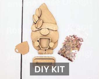 May include: A light brown wooden DIY kit featuring a gnome figure with coffee bean accents on the hat, a separate coffee cup cutout, a rectangular base with a slot, and a small clear plastic bag filled with assorted brown and tan decorative beads.