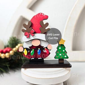 May include: A wooden Christmas gnome with a red hat and antlers, wearing a string of colourful lights. A small green Christmas tree with a yellow star sits beside the gnome. The base is dark brown.