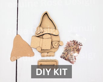 May include: A wooden DIY kit featuring a gnome figure holding a book and a mug, a separate hat piece, and a small plastic bag filled with assorted brown and tan beads. The gnome has a tall hat, a round nose, and a base with a slot for assembly.