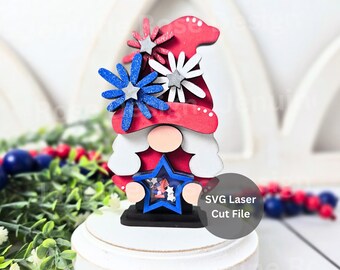 May include: A layered wooden gnome decoration featuring a red hat adorned with blue and white star-shaped flowers. The gnome holds a blue star-shaped shaker filled with small patriotic confetti. The text SVG Laser Cut File is visible on the right.