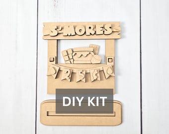 May include: A wooden DIY kit featuring a s'mores design. The kit includes a frame with the word "S'MORES" at the top, a banner with numbers, and a base. The image also includes the text "DIY KIT".