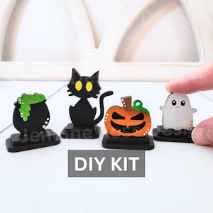 May include: DIY Halloween craft kit with four wooden figures: a black cat, a cauldron with green slime, a pumpkin with a scary face, and a white ghost. The figures are on black bases and the text "DIY KIT" is printed below.