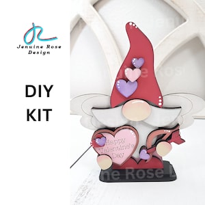 May include: A wooden DIY kit featuring a Valentine's Day gnome. The gnome has a red hat, a white beard, and a heart-shaped body with the text "Happy Valentine's Day." The kit includes a wooden base and decorative hearts.