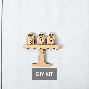 May include: A wooden DIY kit featuring a tiered cake stand with three cupcake-shaped decorations spelling "BOO". The cupcakes have carved details and are placed on a decorative stand. The words "DIY KIT" are in a gray rectangle below the stand.
