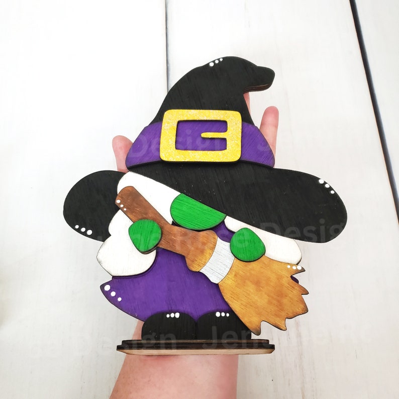 May include: A wooden witch gnome decoration with a black hat, purple dress, green accents, and a brown broom. The gnome is standing on a wooden base.