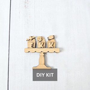 May include: Wooden DIY kit featuring a tiered cake stand with three cupcake toppers spelling out "EKK". Each cupcake is decorated with a bat, skull, and spider. The text "DIY KIT" is displayed below the stand.