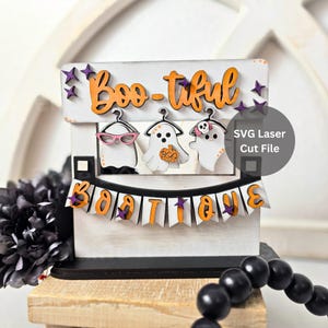 以下が含まれることがあります： 「Boo-tiful」と「Boo-tique」というオレンジ色の文字が書かれた白い木製のハロウィーンの装飾。3体の幽霊の姿が文字の下にぶら下がっています。幽霊はメガネをかけ、カボチャを持ち、頭蓋骨を身につけています。紫色の星が散らばっています。
