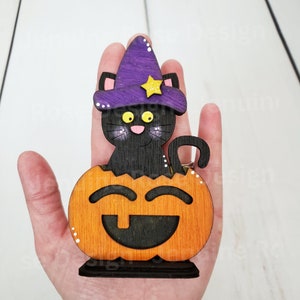 May include: A black cat wearing a purple witch hat with a yellow star sits inside a carved wooden pumpkin with a smiling face.