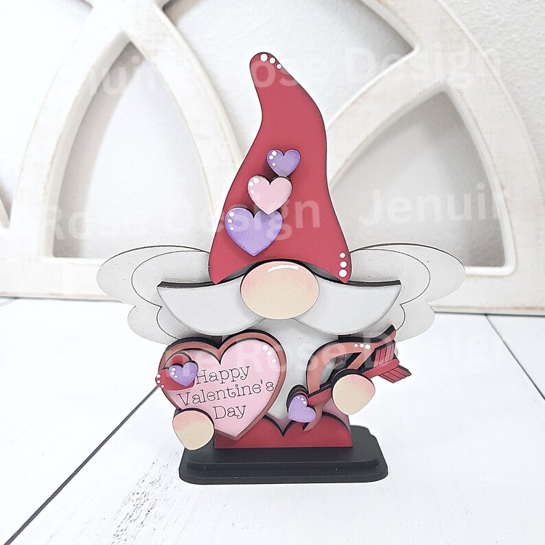 May include: A Valentine's Day gnome figurine with a red hat adorned with three hearts. The gnome holds a heart-shaped sign that reads "Happy Valentine's Day" and an arrow. The base is black, and the overall design is whimsical.