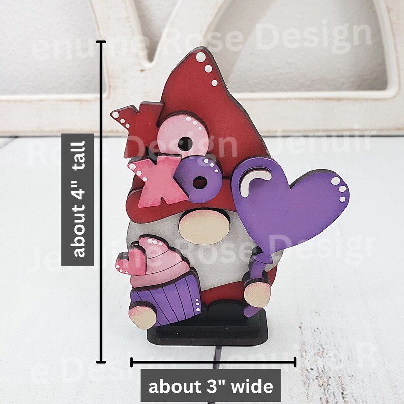 Puede incluir: Una figura de gnomo de madera pintada en rojo, blanco y morado. El gnomo sostiene un globo morado en forma de coraz&oacute;n y un cupcake con un coraz&oacute;n rosa en la parte superior. El gnomo lleva un sombrero rojo con lunares blancos y tiene una barba blanca. El gnomo mide aproximadamente 10 cm de alto y 7,5 cm de ancho. El gnomo dice "XO" en su pecho.
