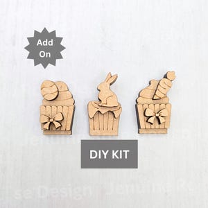May include: Three unfinished wooden cupcake decorations with different Easter themes. The first cupcake has an egg on top, the second has a bunny, and the third has a carrot. The text "DIY KIT" is printed below the cupcakes.