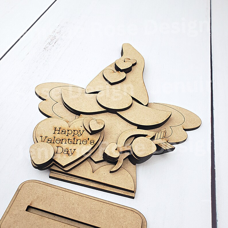 May include: A wooden gnome-shaped Valentine's Day decoration. The gnome has a large hat with heart cutouts, a heart-shaped beard, and holds a heart with an arrow. The text "Happy Valentine's Day" is on a heart. Includes a base.