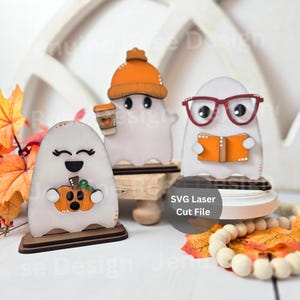 May include: Three wooden ghost figurines with autumnal designs. One ghost holds a pumpkin, another a coffee cup, and the third reads a book. Each ghost is white with black eyes and sits on a wooden base. The text "SVG Laser Cut File" is also visible.