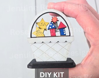 May include: A small wooden basket-shaped decoration featuring two yellow stars with the text USA and est. 1776, alongside three red, white, and blue rocket-shaped cutouts. The piece sits on a black rectangular base and is labeled as a DIY KIT.