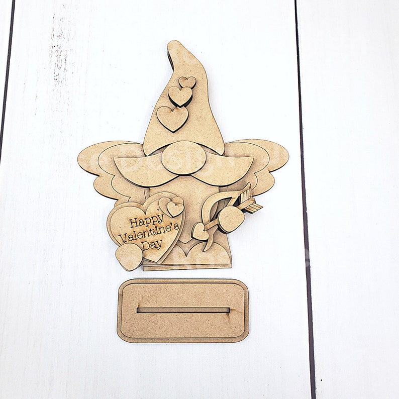 May include: A wooden Valentine's Day gnome decoration. The gnome has a long hat with heart cutouts, a beard, and holds a heart-shaped sign that reads "Happy Valentine's Day." An arrow pierces a heart. Includes a base.