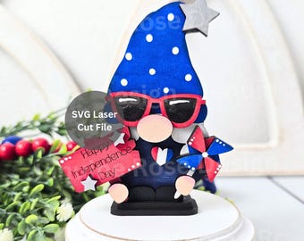 May include: A wooden gnome figurine featuring a blue hat with white polka dots, a silver star, and red-rimmed sunglasses. The gnome holds a red sign reading Happy Independence Day and a red, white, and blue pinwheel, with a heart on its chest.