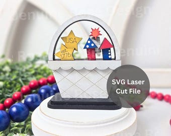 May include: A white wooden basket-shaped tiered tray decor piece featuring two gold stars with the text USA and est. 1776, alongside three red, white, and blue firework-shaped cutouts. The item sits on a round white base with a black bottom layer.