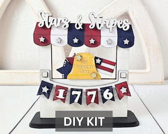 May include: A wooden DIY kit featuring a stand with a red, white, and blue striped awning, the text Stars & Stripes, a yellow bell with the text Let Freedom Ring, and a banner displaying the number 1776 with stars on red and blue pennants.