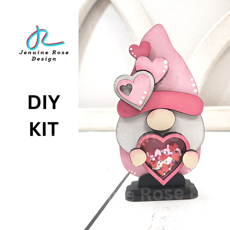 Op de afbeelding: DIY-kit voor een Valentijnsdag-gnome. De gnome is gemaakt van hout en roze, grijs en wit geverfd. Hij heeft een roze hoed met witte stippen en een hartvormig raam met rood en wit confetti erin. De gnome houdt een groot roze hart vast.