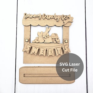 May include: A laser-cut wooden sign featuring the text "Bunny Candy Co." above a window with bunny figures and a banner that reads "SWEETS." The sign includes a slot for display and the text "SVG Laser Cut File."