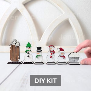 May include: A DIY kit featuring miniature wooden snowmen with various accessories, including a sled and a snowball stand. The snowmen wear hats and scarves in green, red, and black. The text "Snowballs 5¢" is visible on the stand.