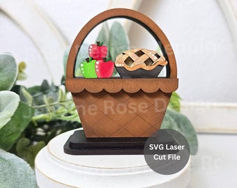 May include: A brown wooden basket-shaped decoration featuring a lattice-patterned base and a rounded handle. Inside the basket are two red apples, one green apple, and a small brown pie with a lattice crust. The text SVG Laser Cut File is visible.