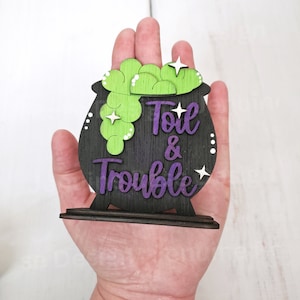 May include: A black cauldron with green and white bubbling potion and the words "Toil & Trouble" in purple lettering. The cauldron is on a wooden stand.