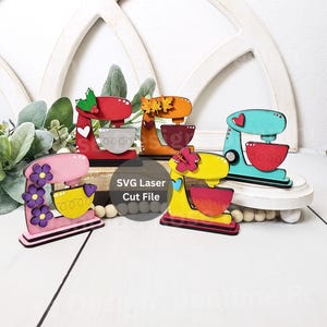 May include: Five wooden stand mixer cutouts in various colours and designs. The stand mixers are decorated with flowers, leaves, and hearts. The text "SVG Laser Cut File" is visible in the image.