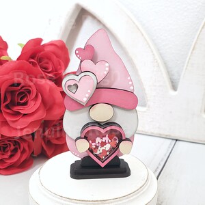 Op de afbeelding: Een roze en witte houten gnome die een hartvormige doos vasthoudt gevuld met rode en roze glitterhartjes. De gnome draagt ​​een roze hoed met witte stippen en heeft een witte baard.