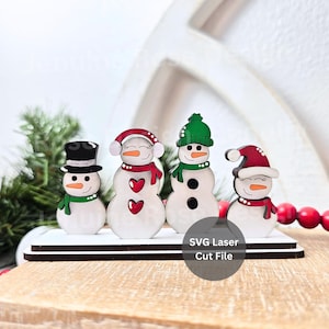 Può includere: Un set di quattro statuette di pupazzi di neve in legno. Ogni pupazzo di neve ha un cappello, una sciarpa e dettagli di bottoni o cuori unici. I pupazzi di neve sono esposti su una base di legno bianca e marrone, a tema natalizio.