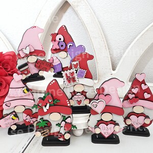 Puede incluir: Un conjunto de ocho gnomos de San Valent&iacute;n, cada uno con un dise&ntilde;o diferente. Los gnomos est&aacute;n hechos de madera y pintados en varios tonos de rojo, rosa y blanco. Presentan corazones, flores y otras decoraciones con tem&aacute;tica de San Valent&iacute;n. Los gnomos est&aacute;n de pie sobre una superficie blanca.