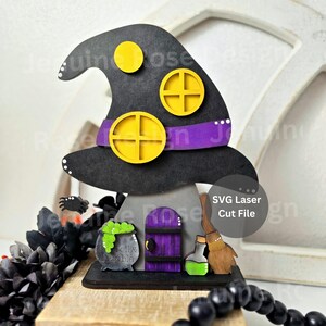May include: A Halloween decoration in the shape of a witch's hat, with yellow circles and a purple band. It includes a cauldron, broom, and door. The text "SVG Laser Cut File" is visible. The design is made for laser cutting.