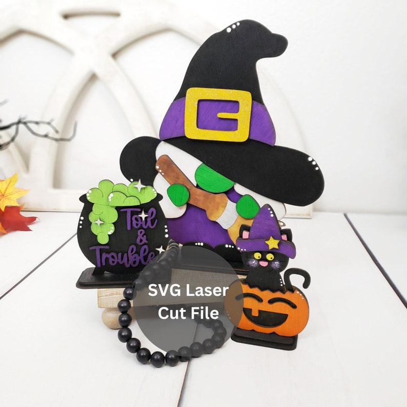 May include: A wooden Halloween witch figurine with a black hat, purple dress, and green broom. The witch is holding a cauldron that says "Toil & Trouble". A black cat with a purple hat sits on a pumpkin. The figurine is on a wooden base with a black beaded bracelet.