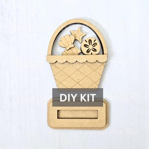 May include: A light brown wooden cutout in the shape of a basket filled with a seashell, a starfish, and a sand dollar. The basket features a cross-hatched pattern and a handle. A rectangular base is attached at the bottom with the text DIY KIT.