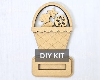 May include: A light brown wooden cutout in the shape of a basket filled with a seashell, a starfish, and a sand dollar. The basket features a cross-hatched pattern and a handle. A rectangular base is attached at the bottom with the text DIY KIT.