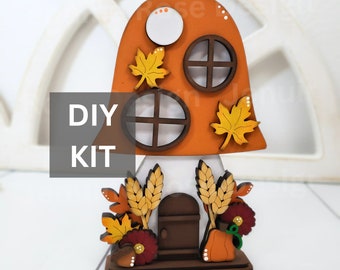 May include: DIY kit for a mushroom house craft project. The kit includes a wooden mushroom shaped house with a white base, a brown door, and windows. The mushroom is decorated with fall leaves and a pumpkin. The kit is labeled "DIY KIT".