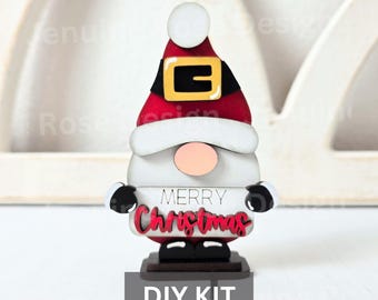 May include: A wooden Christmas gnome DIY kit. The gnome has a red hat with a black band and gold buckle, a white beard, and a pink nose. The words "MERRY Christmas" are displayed on the front. The base of the gnome is brown, and the text "DIY KIT" is at the bottom.