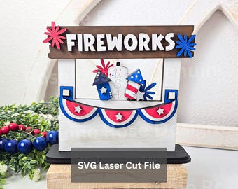 May include: A tiered tray sign featuring a brown wooden top board with the white text FIREWORKS, accented by red and blue starbursts. The white base displays a red, white, and blue bunting with stars and miniature rocket cutouts labeled USA.