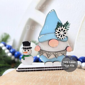 May include: A wooden winter-themed decoration featuring a gnome and a snowman. The gnome has a light blue hat and a banner that says "BAR". The snowman wears a black top hat and a green scarf. A snowflake and text "SVG Laser Cut File" are also visible.