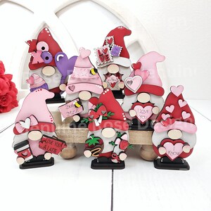 Puede incluir: Ocho gnomos de San Valent&iacute;n con diferentes dise&ntilde;os y lemas. Los gnomos est&aacute;n hechos de madera y pintados en tonos de rojo, rosa y blanco. Algunos de los lemas incluyen "Te amo", "S&eacute; m&iacute;o" y "Te amo un latte".